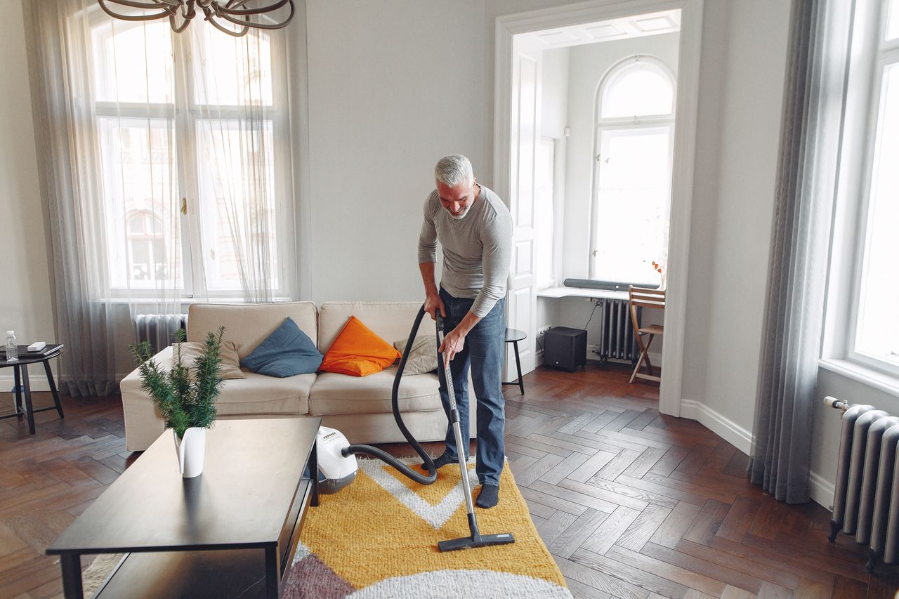 Vue de dessus d’un homme barbu aux cheveux gris, concentré, en train d’aspirer un tapis à la maison